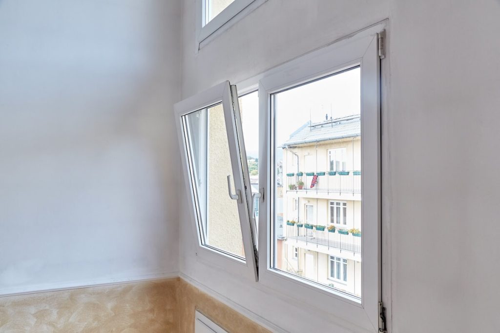 A modern white-framed tilt-and-turn window, partially open, in a bright interior. Through the window, a beige apartment building with balconies and flower boxes is visible.